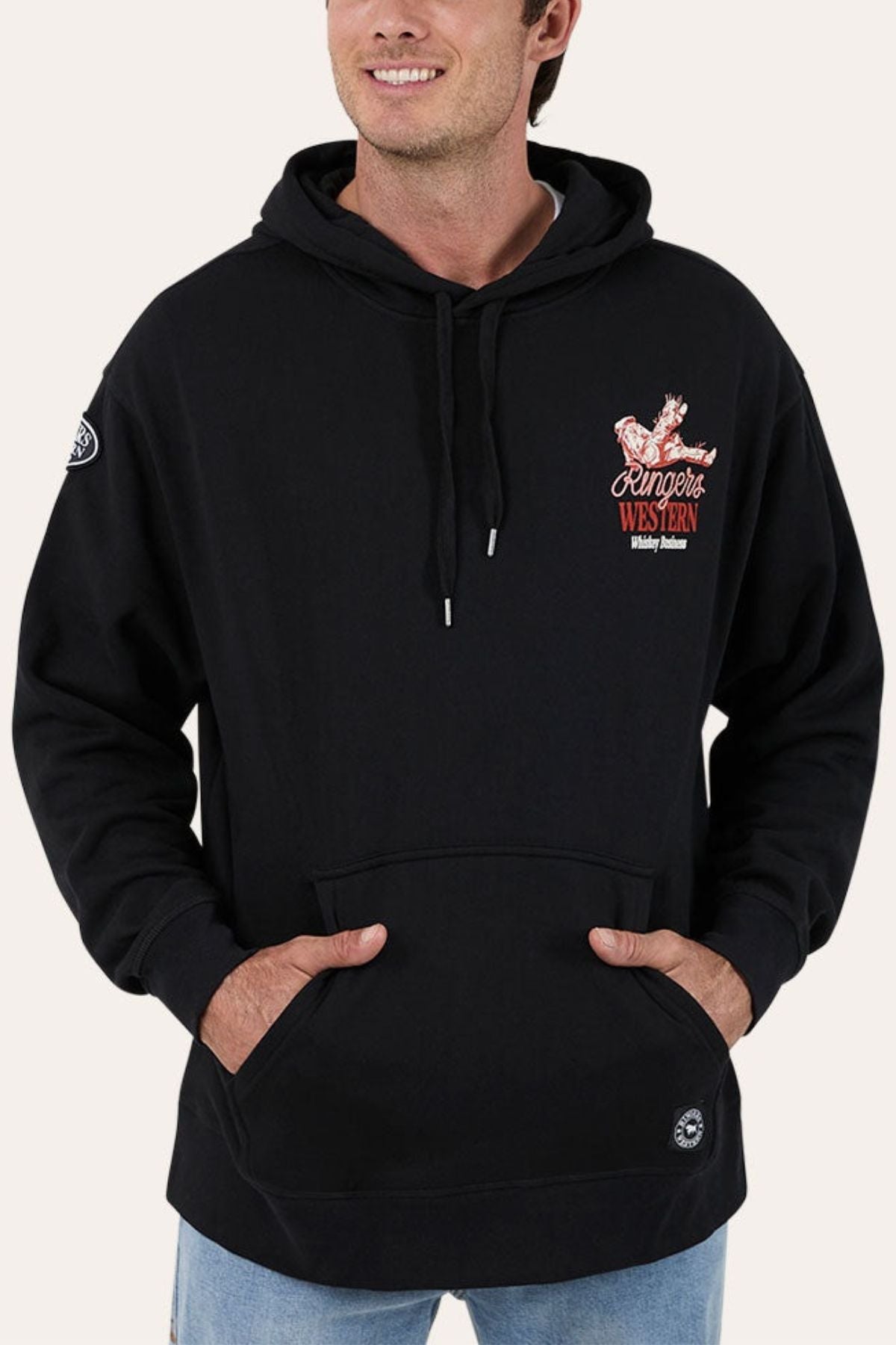 A man smiles with his hands in the pocket of a black Ringers Western Whiskey Business Pullover Hoodie, which has brushed fleece, a front pocket, and a small red and white embroidered chest logo.