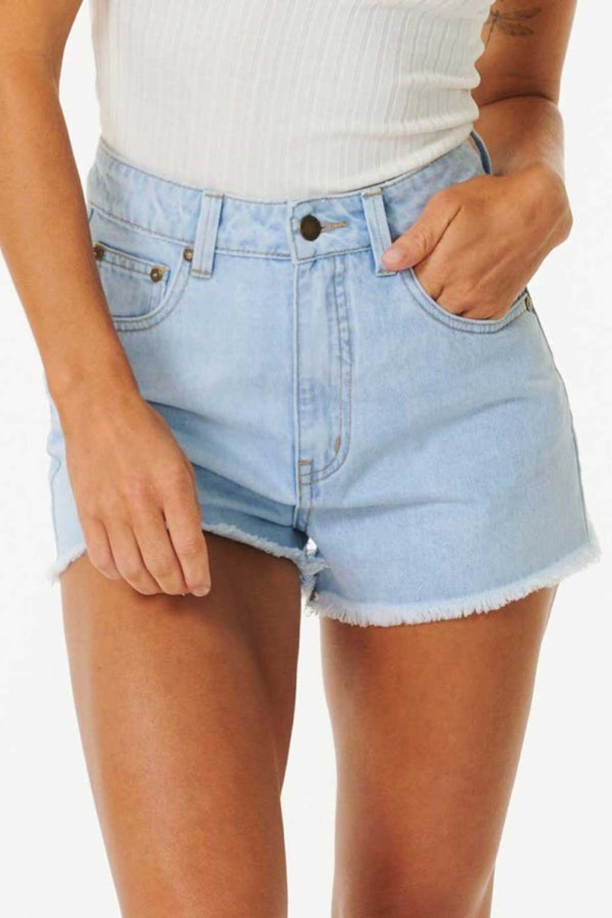 A person dressed in Rip Curl Amy High Waist Shorts, featuring a frayed hem and light blue denim, paired with a white ribbed tank top stands with hands in pockets, ready for summer adventures against a plain white background.