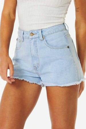 The individual is sporting the Rip Curl Amy High Waist Short in a light blue shade, featuring a frayed hem for a casual look, paired with a white ribbed tank top. Ideal for summer adventures, these denim shorts include a button closure and front pockets. The backdrop is light and neutral.