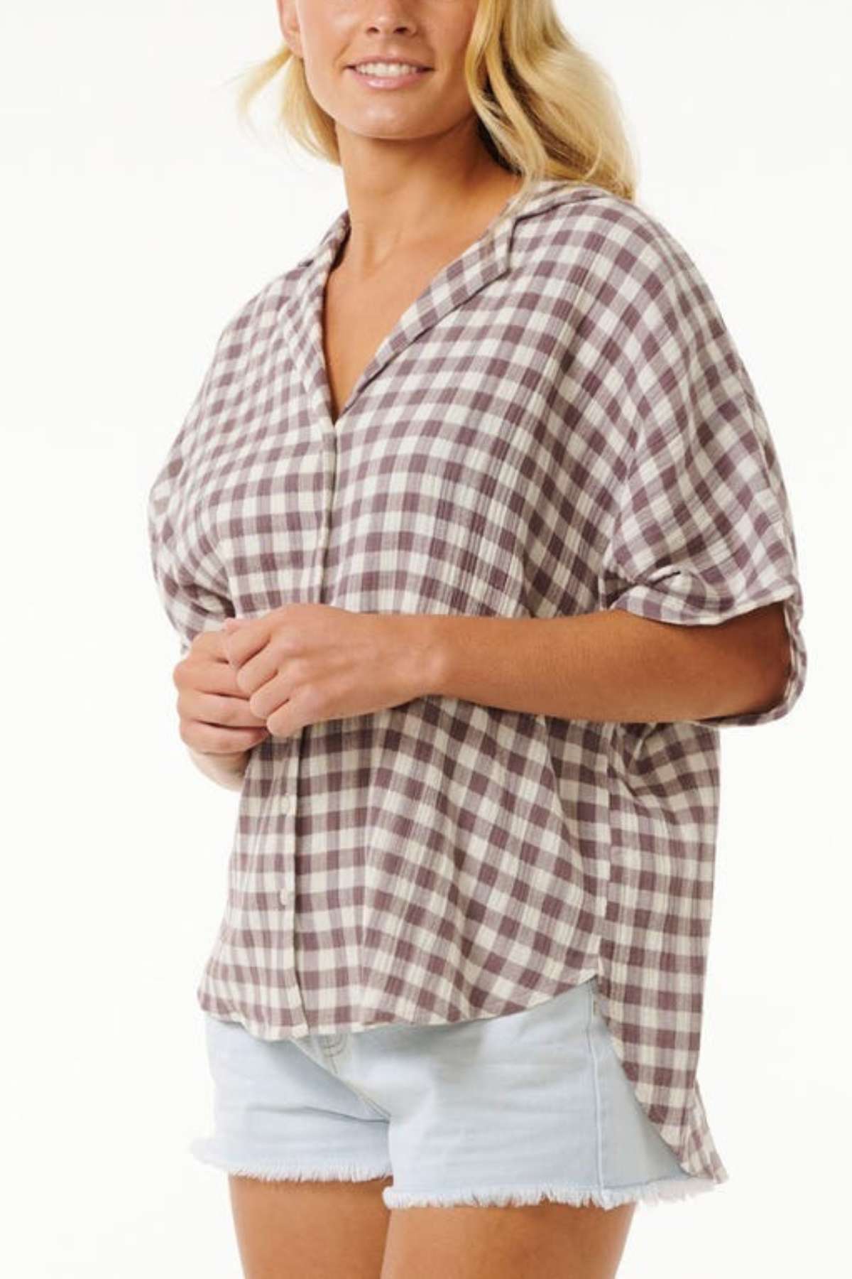 A person wearing a loose-fitting, short-sleeved checkered shirt in brown and white over light blue denim shorts stands against a plain background. The Rip Curl Premium Surf Check Shirt is untucked, and the shorts have frayed hems. Perfect for the ocean lover, this look is effortlessly casual.