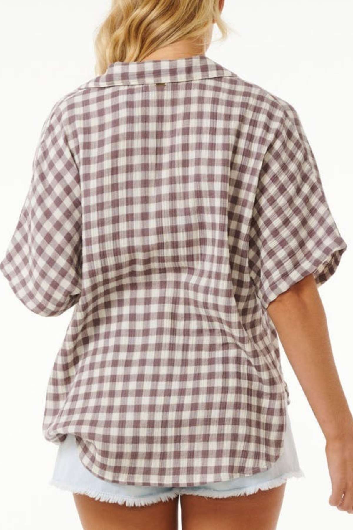 A person with wavy blonde hair is wearing a loose-fitting, short-sleeved Rip Curl Premium Surf Check Shirt in a brown and white plaid pattern. They're paired with light blue denim shorts featuring frayed edges. Shot from behind against a plain background, this ocean lover's style exudes casual charm.