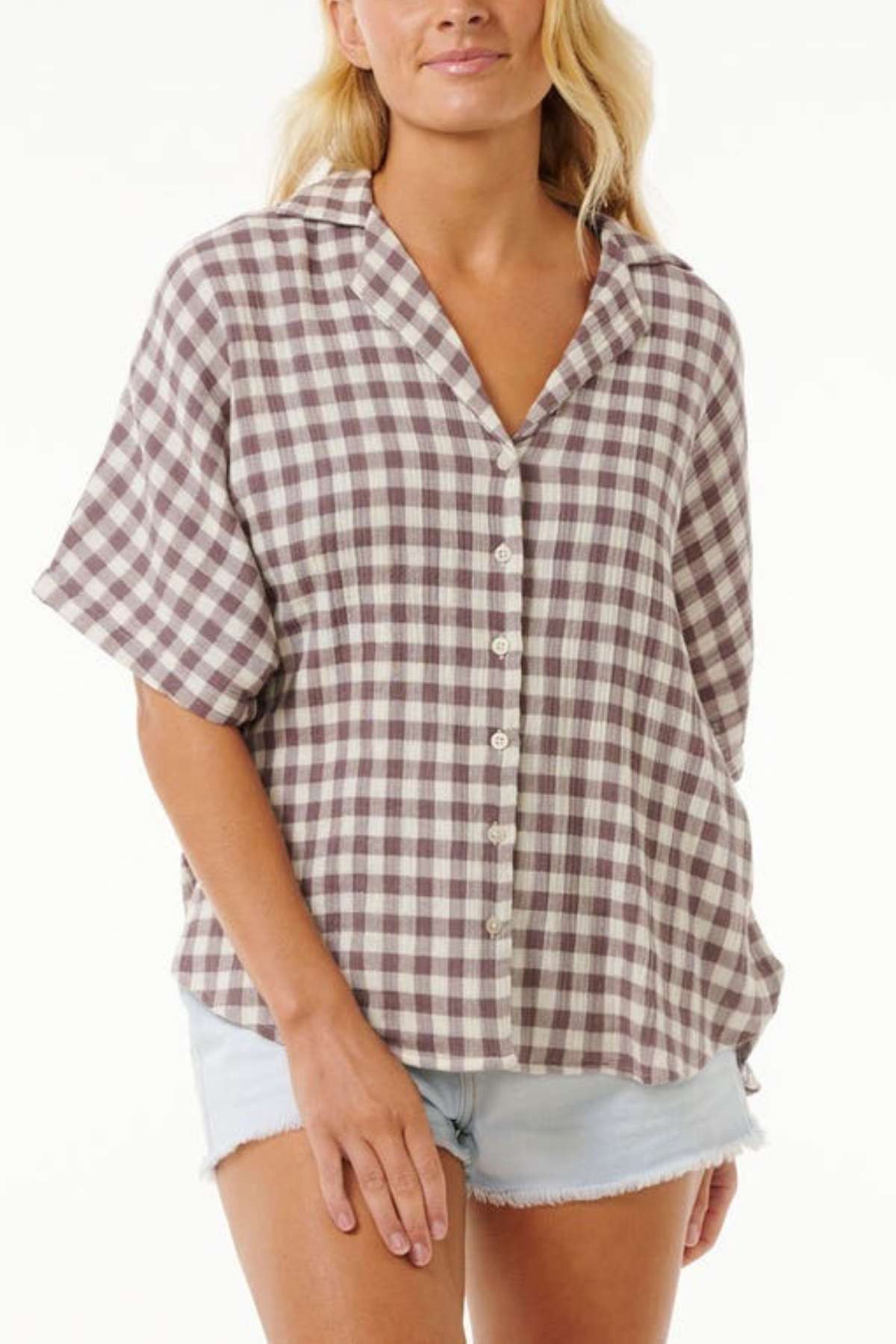 The woman is wearing a short-sleeved, brown and white checkered Rip Curl Premium Surf Check Shirt made from 100% cotton. Paired with her light-colored denim shorts, her style exudes the casual vibes of an ocean lover against the plain white backdrop.