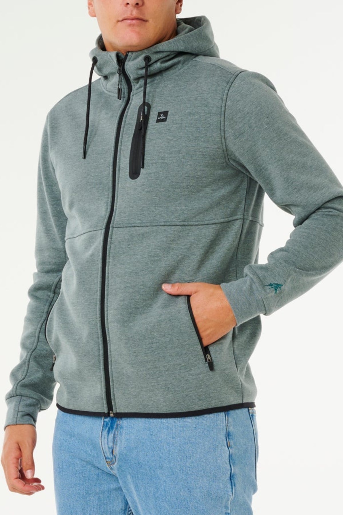 Wearing a light gray Rip Curl Anti Series Departed Zip Thru hoodie with black drawstrings and a hood, paired with light blue jeans. One hand is in the pocket, the other relaxed at their side. The face is partially out of frame.