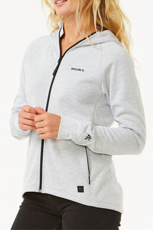 A woman wearing a light gray Rip Curl Anti Series Flux III Jacket with a hood, black zipper, and chest logo stands against a white background. This lightweight, shower-resistant jacket offers both style and function.