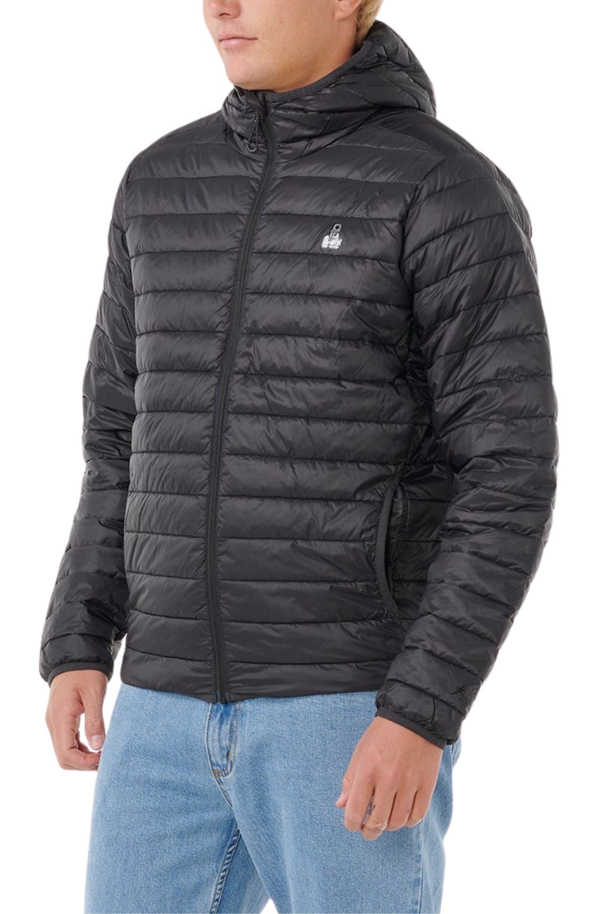 Someone wearing the Rip Curl Anti Series Search Puffer Hood, a black, packable puffer jacket with horizontal quilting, paired with light blue jeans, stands against a white background.