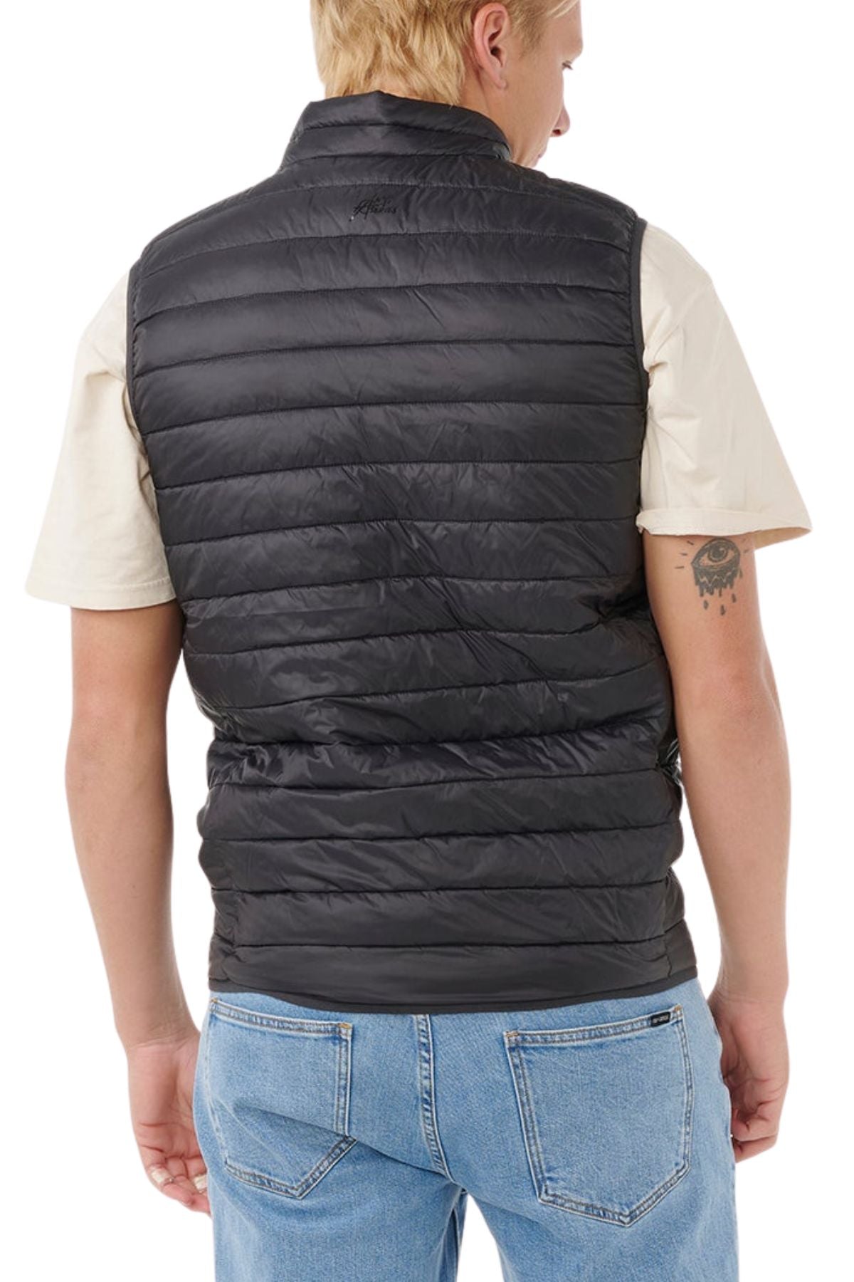 Someone with short blonde hair and a tattoo on their left arm is shown from the back, wearing the Rip Curl Anti Series Search Puffer Vest in black over a beige short-sleeve shirt and blue jeans. The vest is water repellent and lightweight.