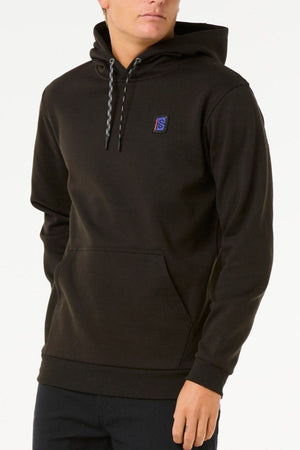 A person wears the Rip Curl Anti Series Search Base Hood, a black water repellent hoodie with a front pocket, patterned drawstrings, and a small rectangular chest logo, standing against a plain white background.