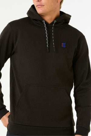 A person wears a black Rip Curl Anti Series Search Base Hood sweatshirt with gray drawstrings, a front pocket, and a small blue "S" patch on the chest. The water-repellent hoodie is set against a plain background.