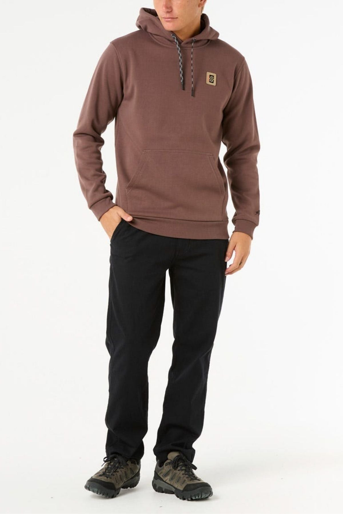 A person wears the Rip Curl Anti Series Search Base Hood, brown bonded polar fleece with a small logo patch, black pants, and brown sneakers, standing with one hand in their pocket against a plain white background.