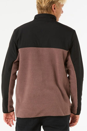 A person with short blond hair is seen from behind, wearing a Rip Curl Anti Series Search Zip Crew featuring a black upper part, textured brown lower section, and water repellent treatment. The background is plain white.
