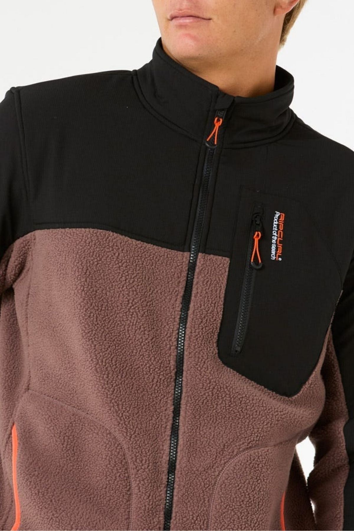 A person wears the Rip Curl Anti Series Search Zip Crew, featuring a black upper, brown polyester polar fleece lower section, zippered chest pocket with orange accents, and visible logo. The person's face is partially out of frame.