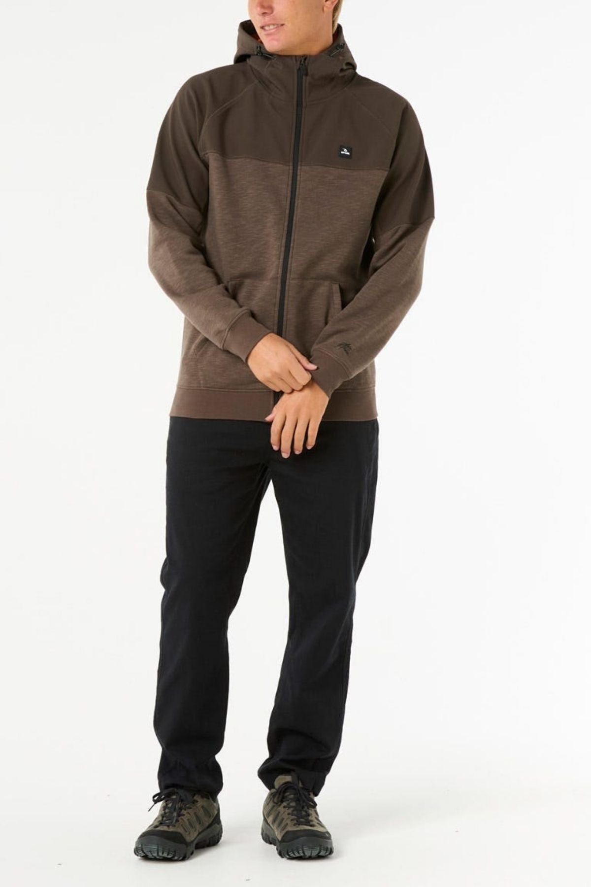 A person stands against a white background, wearing the Rip Curl Anti Series Viral Zip Thru jacket in brown with black pants and dark green sneakers, hands near the waist and face partially turned away, showcasing stylish adventure wear.