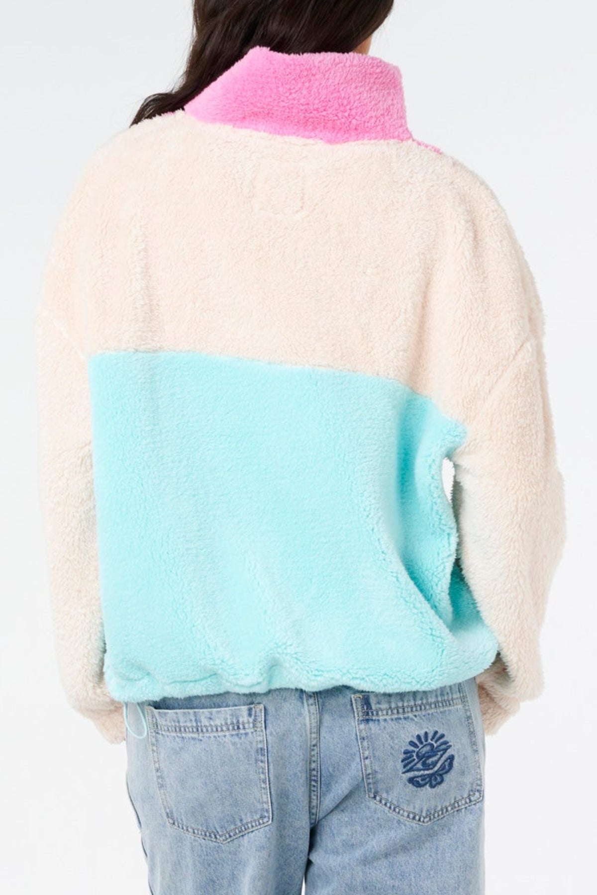 A person is seen from the back in a Rip Curl Block Party II Zip Through Polar Fleece, featuring a pink collar, cream top, and light blue bottom, styled with light blue jeans against a plain background.