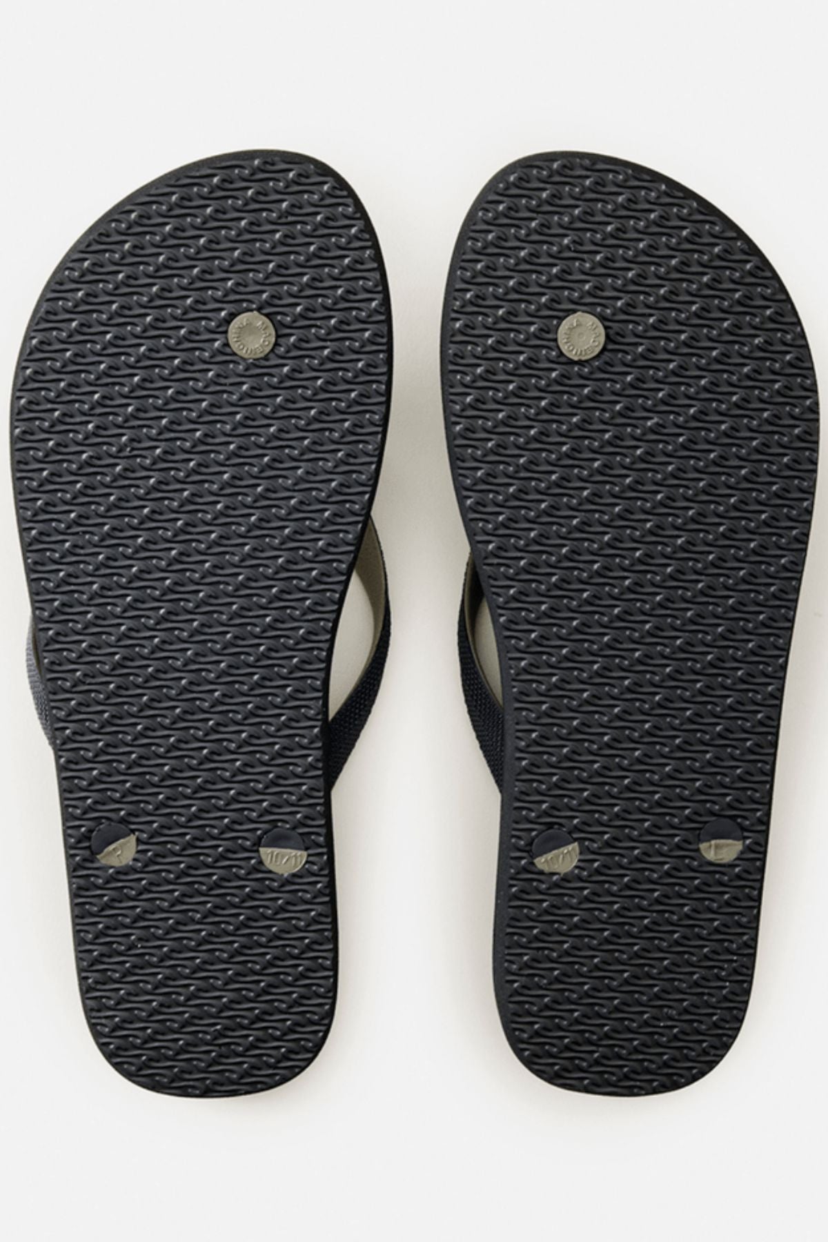 A pair of black Rip Curl Camo Bloom Thongs shown from the bottom, highlighting textured soles and the circular and oval plugs where straps attach. Crafted with BLOOM algae foam for comfort.