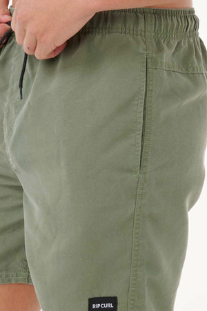 Close-up of a person in Rip Curl Bondi Volley shorts in olive green, featuring an elastic waistband, side pocket, and a small "RIP CURL" label near the hem. Made from recycled PET. The left hand rests by the waistband.