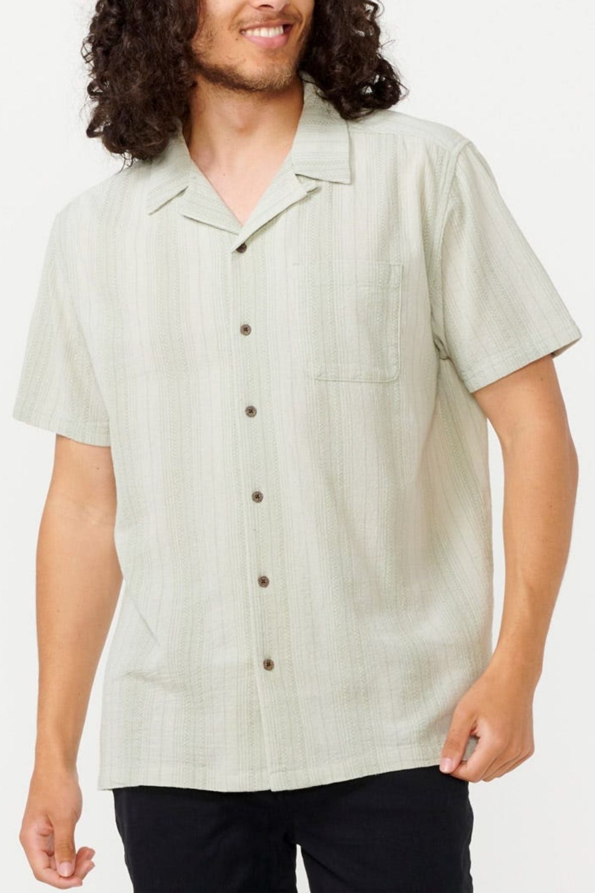 A person with curly hair wears a Rip Curl Check Mate Short Sleeve Shirt in light green, crafted from cotton gauze with subtle stripes. Featuring a chest pocket and dark buttons, it's styled elegantly with dark pants against a plain white background.