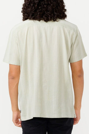 A person with curly hair is seen from the back, wearing the light-colored Rip Curl Check Mate Short Sleeve shirt and dark pants, against a plain white background.