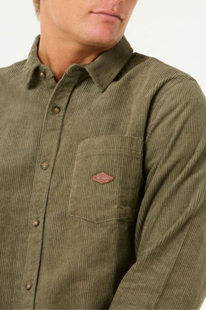 A person wearing a Rip Curl Classic Surf Cord Shirt in olive green cotton corduroy with a chest pocket and small brown patch; their face is partially out of frame.