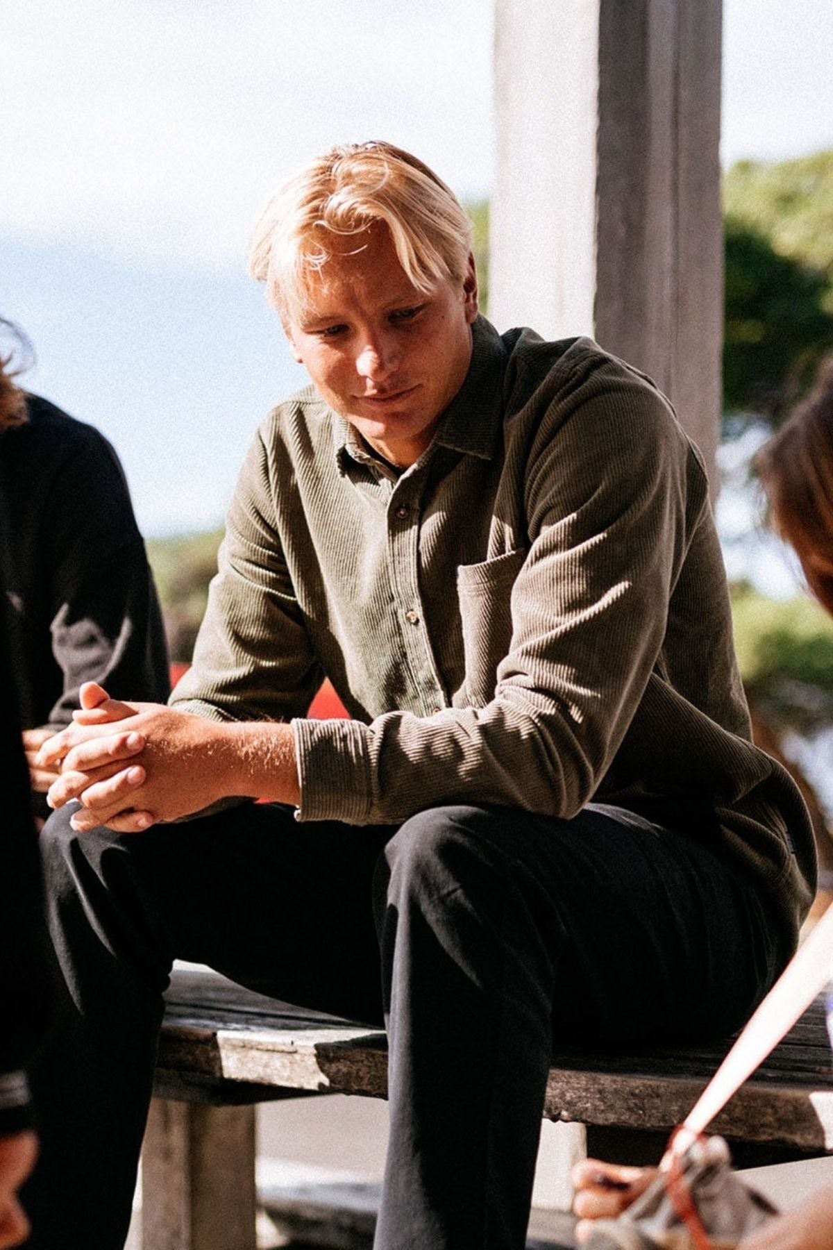 A blond man in a green Rip Curl Classic Surf Cord Shirt and black pants sits thoughtfully on an outdoor wooden bench, hands clasped and sunlight highlighting his face.