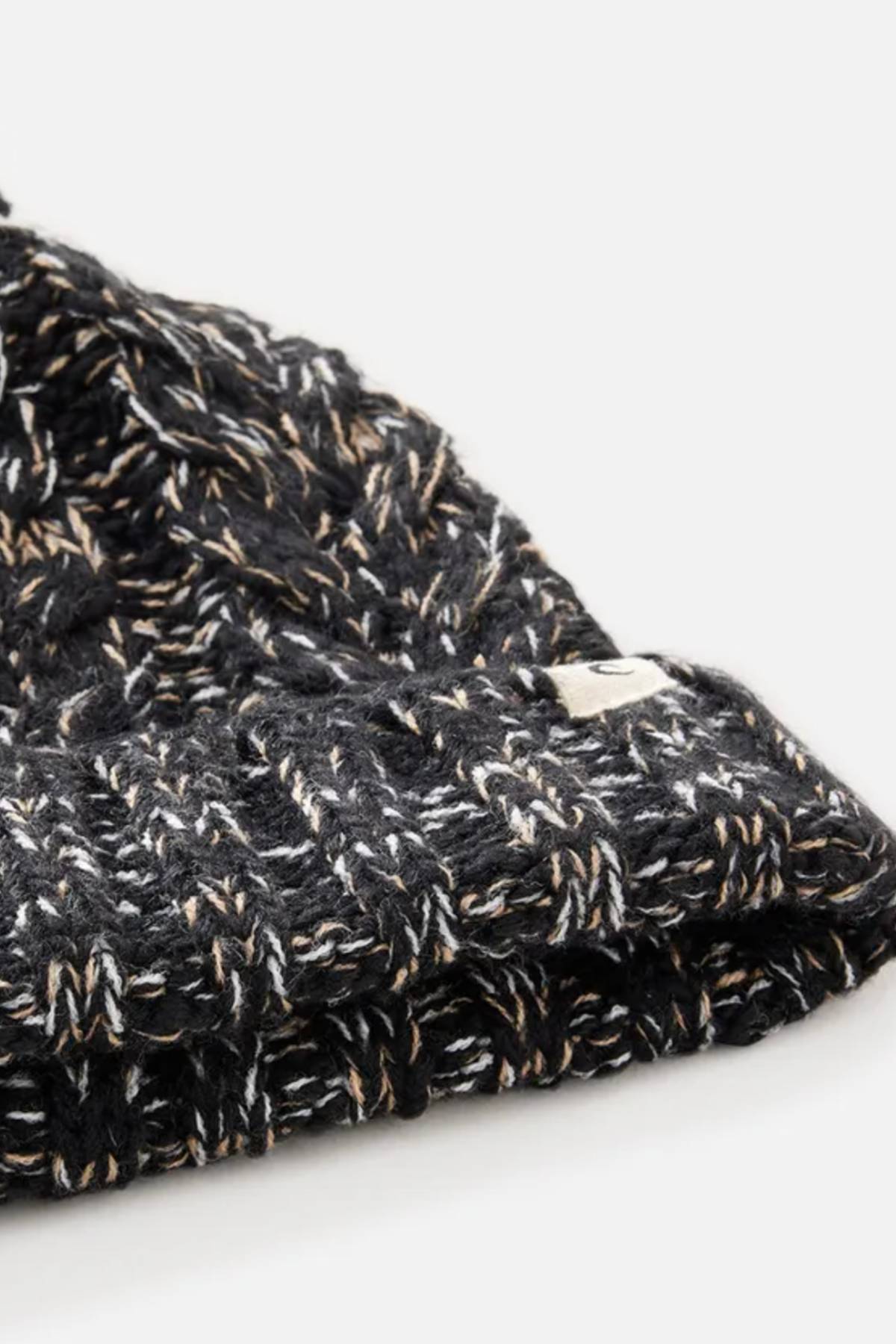 Close-up of the Rip Curl Classic Regular Pom Pom Beanie, a black knit with white and tan threads, ribbed texture, small white edge tag, and soft acrylic yarn. Shown against a plain light background.