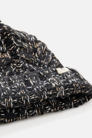 Close-up of the Rip Curl Classic Regular Pom Pom Beanie, a black knit with white and tan threads, ribbed texture, small white edge tag, and soft acrylic yarn. Shown against a plain light background.