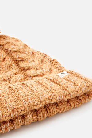 Close-up of the Rip Curl Classic Regular Pom Pom Beanie, a knitted mustard yellow hat with a ribbed edge, made from soft acrylic yarn and featuring a small white tag on the brim, set against a plain light background.