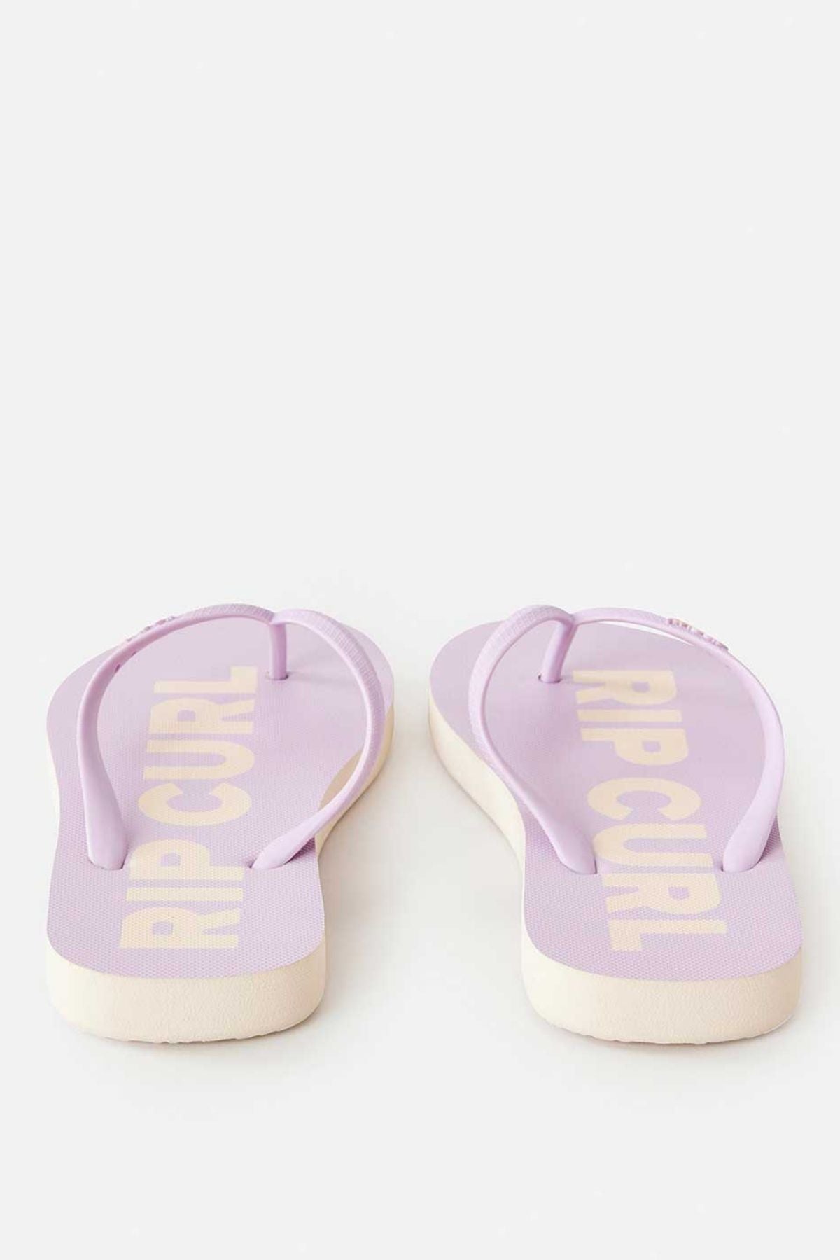 The Rip Curl Classic Surf Bloom Thongs in lavender feature white soles and prominently display the "RIP CURL" logo on the footbed. These eco-friendly flip flops are made from BLOOM algae-blended foam, and are positioned toe-side away on a light background.