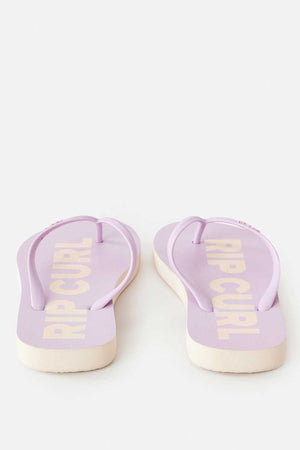 The Rip Curl Classic Surf Bloom Thongs in lavender feature white soles and prominently display the "RIP CURL" logo on the footbed. These eco-friendly flip flops are made from BLOOM algae-blended foam, and are positioned toe-side away on a light background.