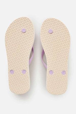 The image displays the bottom view of a pair of Rip Curl Classic Surf Bloom Thongs in beige, featuring BLOOM algae-blended foam for an eco-conscious design. The textured soles are complemented by subtle lavender accents, with one thong marked as size "5/6" and the other labeled "L." These sustainable flip-flops are elegantly positioned on a white background.