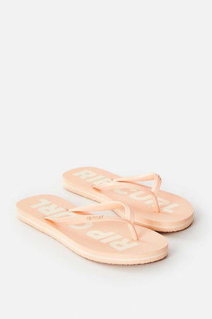 The Rip Curl Classic Surf Bloom Thongs come in a light peach shade featuring white abstract lettering on the footbed. Made with eco-friendly BLOOM™ algae-blended foam EVA for sustainable comfort, they boast minimalist thong straps and are showcased on a light gray background.