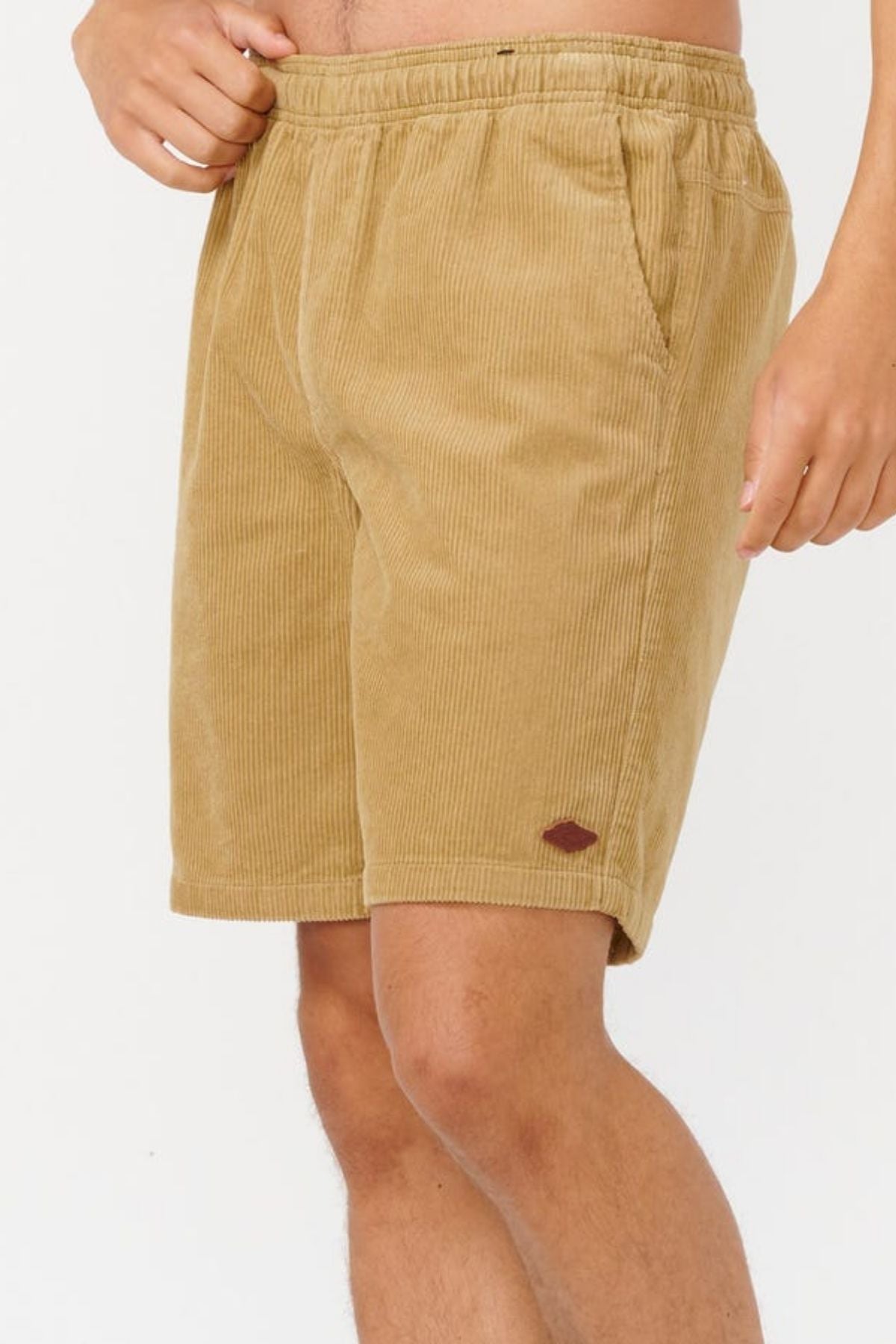 A person wears Rip Curl Classic Surf Cord Volley shorts—OG fit tan cotton corduroy with an elastic waistband, side pockets, and a small brown patch above the left hem. Ideal for beach casual wear. The background is plain white.