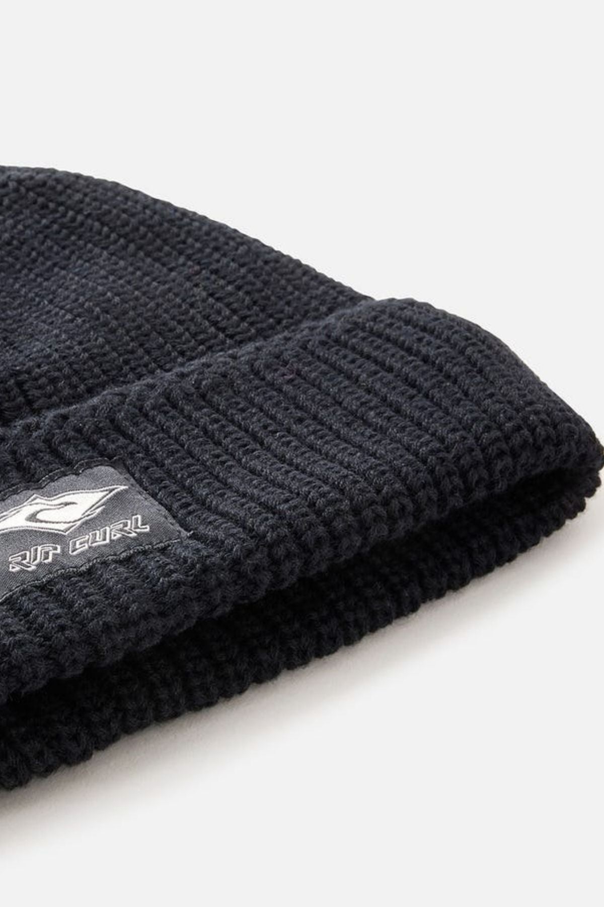 Close-up of the Rip Curl Classic Surf Lined Reg Beanie in black, featuring a folded cuff with a small rectangular black and white Rip Curl logo tag. The background is plain and light-colored.