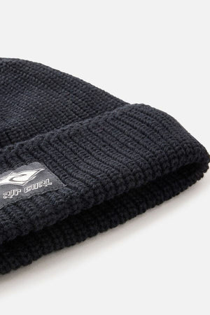 Close-up of the Rip Curl Classic Surf Lined Reg Beanie in black, featuring a folded cuff with a small rectangular black and white Rip Curl logo tag. The background is plain and light-colored.