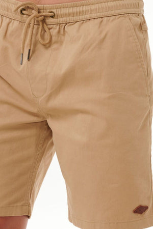 A person wears Rip Curl Classic Surf Volley tan cotton shorts with an elastic waistband and adjustable drawcord for a flexible fit. The shorts display a small stitched logo on the lower left leg, set against a plain, blurred background.