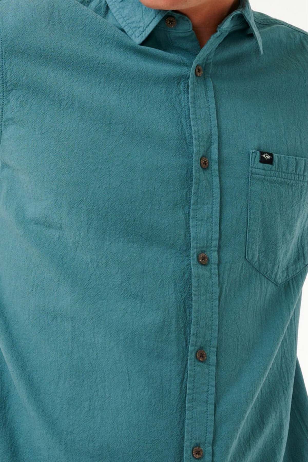 A close-up shows someone wearing the Rip Curl Classic Surf Washed Short Sleeve Shirt in teal, featuring a chest pocket with a small logo tag. The shirt is made from cotton gauze fabric with an enzyme wash finish and is accented with brown buttons. The person's face is not visible.