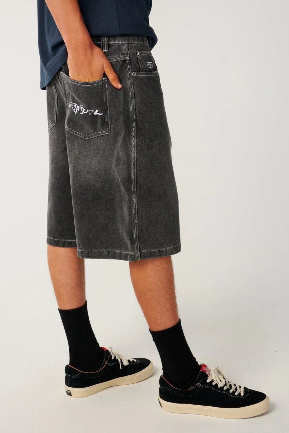 Person wearing relaxed baggy Rip Curl Archive Dirty Denim Walkie Walkshorts with a graphic on the back pocket, paired with black sneakers and black socks.