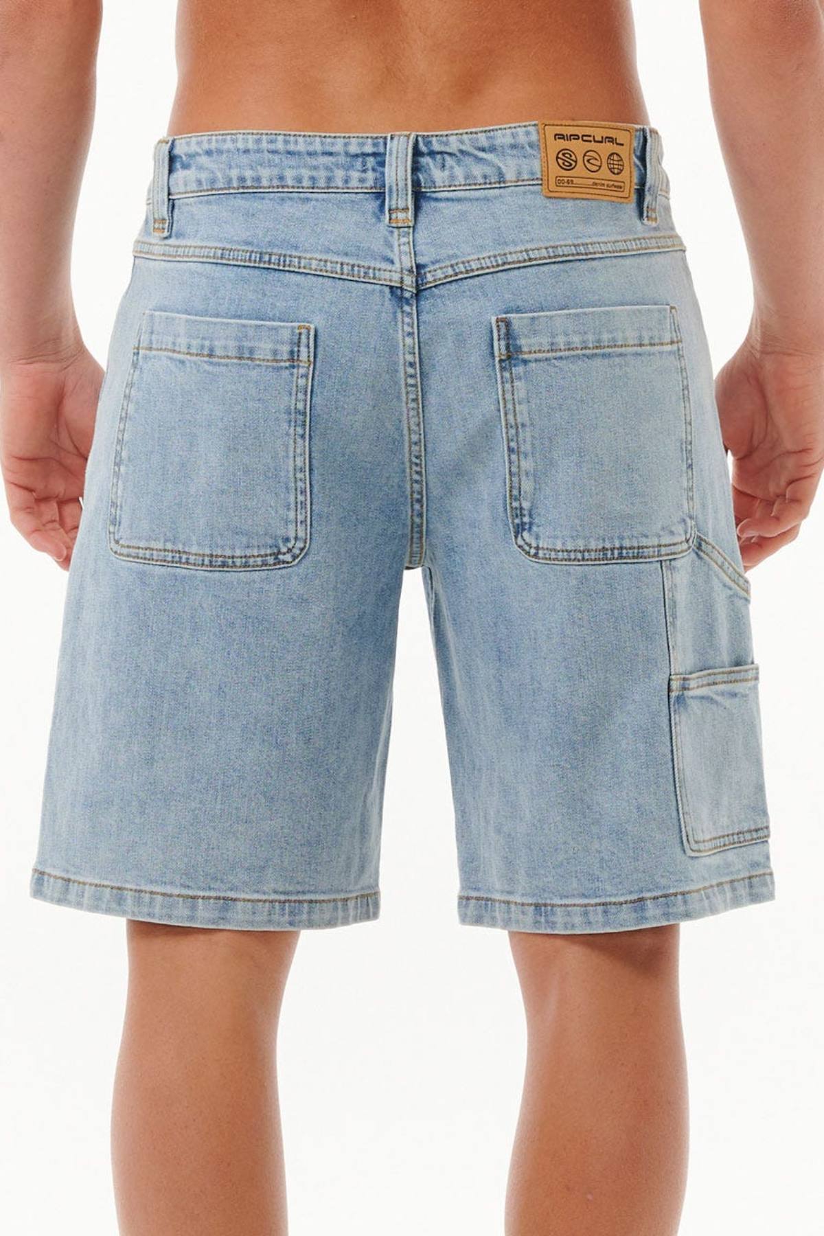 Seen from the back, a person wears the Rip Curl Dosed Denim Walkshort in light blue, styled with large rear pockets and a tan Rip Curl patch on the waistband for a classic 90s look.