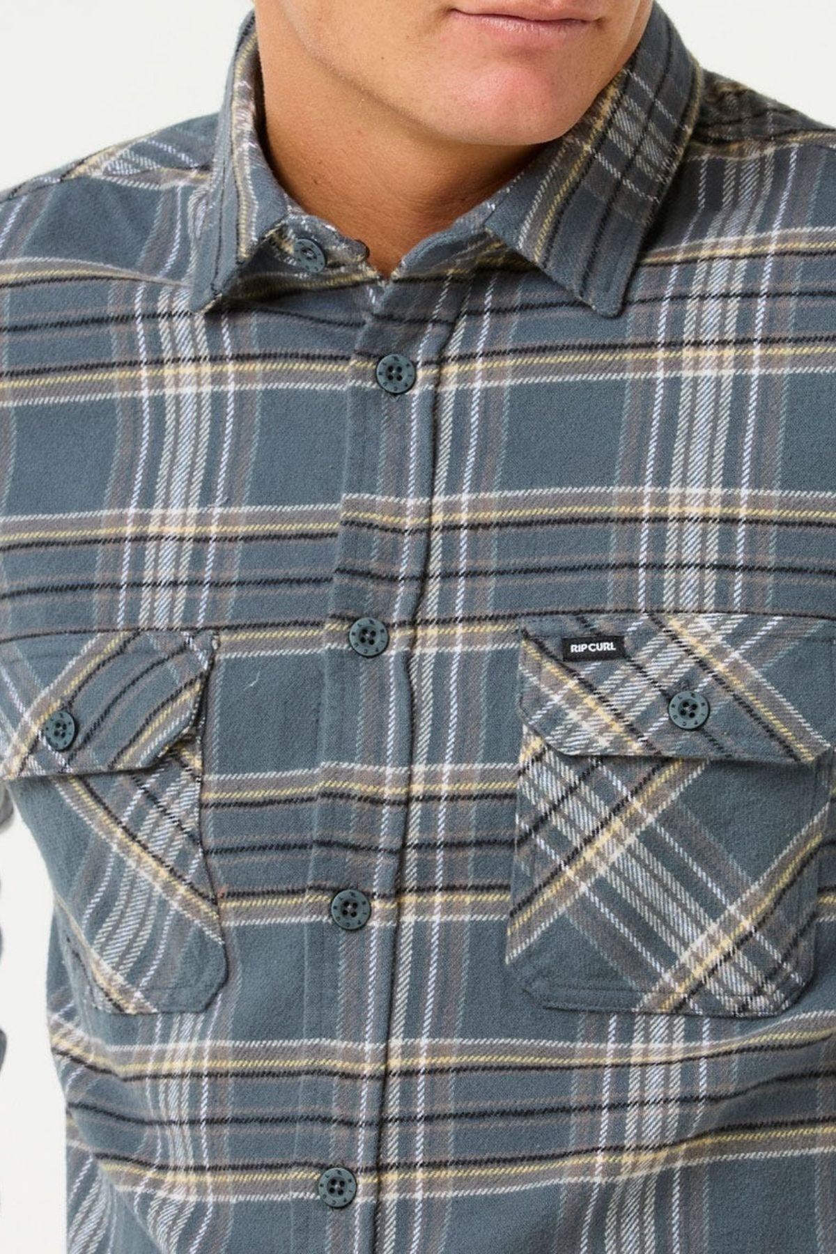 A person wearing the Rip Curl Griffin Flannel Shirt in blue and gray with buttoned chest pockets, including a visible "Rip Curl" label. The photo is cropped to show just the lower face and upper torso.