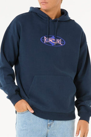 A person wears the Rip Curl Heritage Oval Hood in dark blue, made from soft knit fabric with a front pocket, paired with light blue jeans, standing against a plain background with one hand in their pocket.
