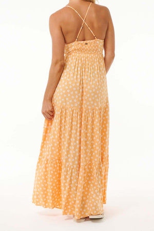The individual is dressed in a long, sleeveless orange dress adorned with white floral designs, made from soft Viscose. This outfit includes a shirred back and thin straps crossing over the shoulders, echoing the style of the Rip Curl High Tide Ditsy Maxi Skirt. They are posing with their back to the camera against a plain backdrop.
