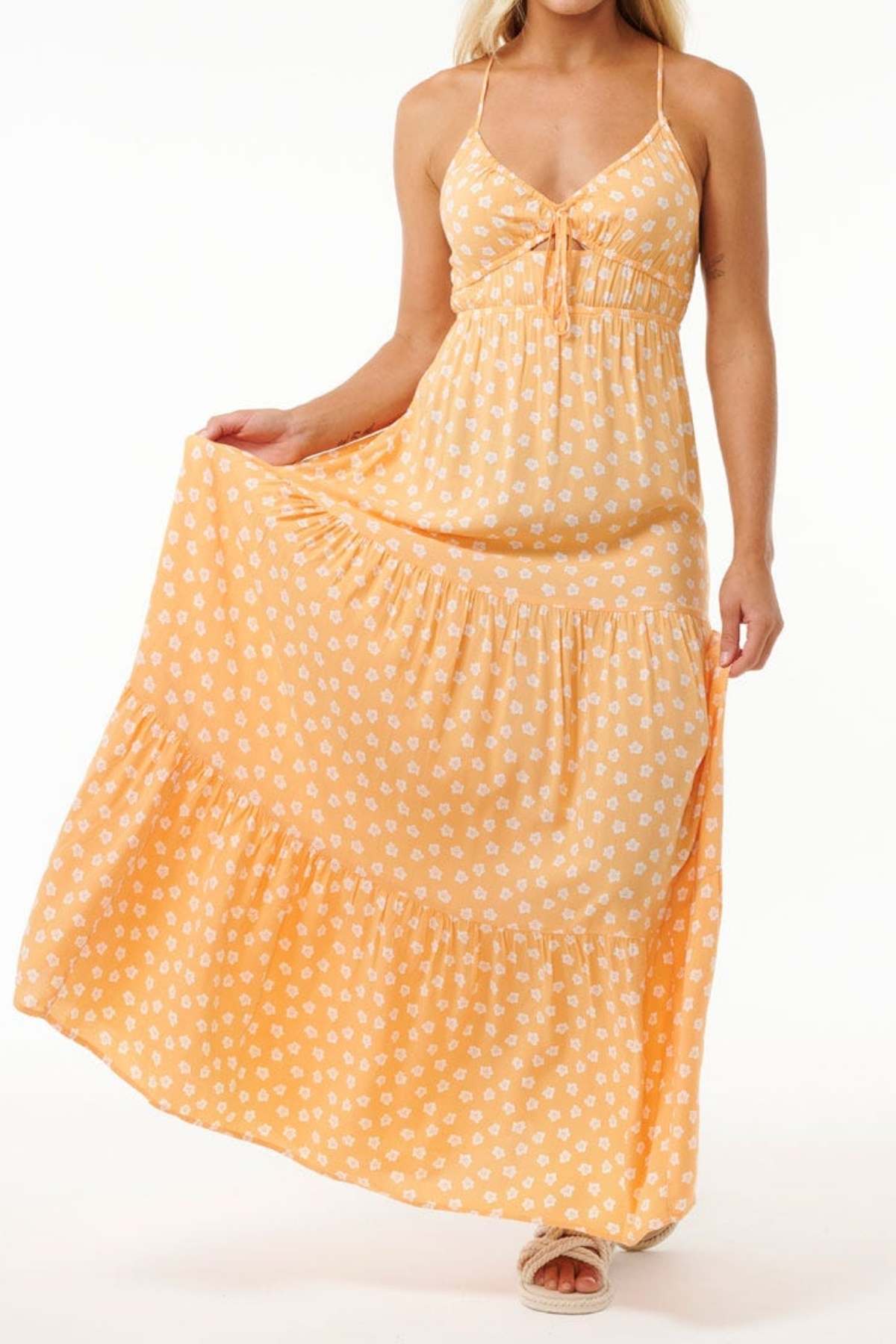 A woman wearing a long, flowing yellow sundress adorned with white floral patterns, similar to the Rip Curl High Tide Ditsy Maxi Skirt, gracefully holds the left side of her dress. She complements her outfit with tan sandals and has light blonde hair. The background is plain white.
