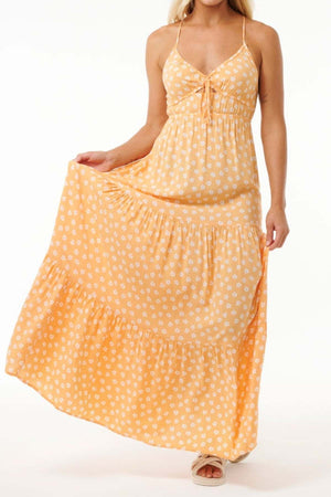 A woman wearing a long, flowing yellow sundress adorned with white floral patterns, similar to the Rip Curl High Tide Ditsy Maxi Skirt, gracefully holds the left side of her dress. She complements her outfit with tan sandals and has light blonde hair. The background is plain white.