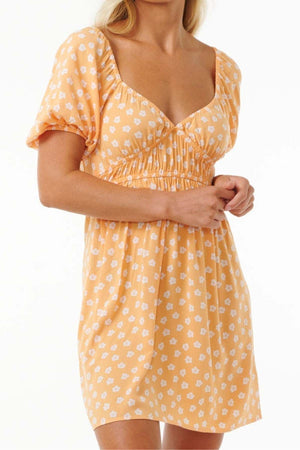 A person is wearing the fashion-forward Rip Curl High Tide Ditsy Mini Dress in an orange floral pattern, featuring a ruched bodice and sweetheart neckline. This Lenzing EcoVero POPLIN piece ends above the knee, with hands held together in front.