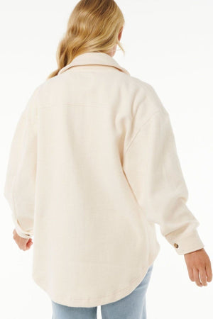 A person with long, wavy blonde hair is seen from behind in a Rip Curl High Tide Fleece Shacket, cream color, paired with light blue jeans—ideal for adventurous people—set against a plain white background.