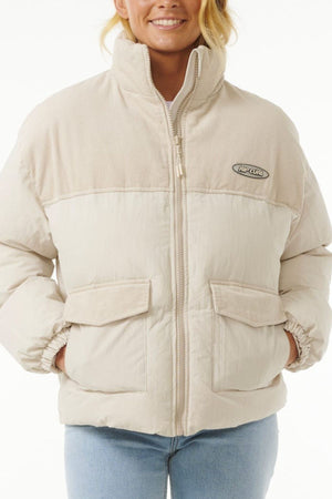 A person wears a light beige Rip Curl High Tide Mixed Cord Puffer jacket with a high collar, two large front pockets, and an embroidered chest logo, paired with light blue jeans against a plain white background.
