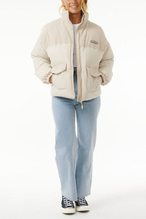 A person wearing the Rip Curl High Tide Mixed Cord Puffer, embroidered logo visible, light blue jeans, and black-and-white sneakers stands facing forward with hands in pockets against a plain white background.