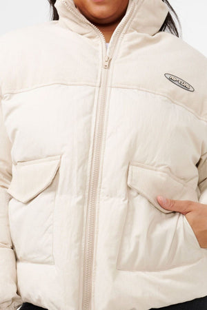 A person wears a cream Rip Curl High Tide Mixed Cord Puffer with front zipper, cotton cord panel, two flap pockets, and a small chest logo. Only part of the face and body are visible.