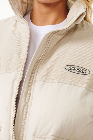 Close-up of a person wearing the Rip Curl High Tide Mixed Cord Puffer in light beige, with an embroidered "RIP CURL" logo on the chest, cotton cord panel details, and layered over a white shirt. Only the lower face and upper torso are shown.