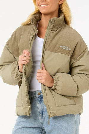 A woman wears the Rip Curl High Tide Mixed Cord Puffer in light beige over a white shirt and light blue jeans. Smiling, she holds the zipper of the jacket, her long blonde hair loose for a relaxed, effortless look.