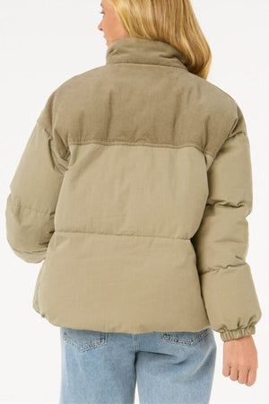 A woman with long blonde hair is seen from behind wearing the Rip Curl High Tide Mixed Cord Puffer with corduroy accents and an embroidered logo, paired with light blue jeans, set against a plain white background.